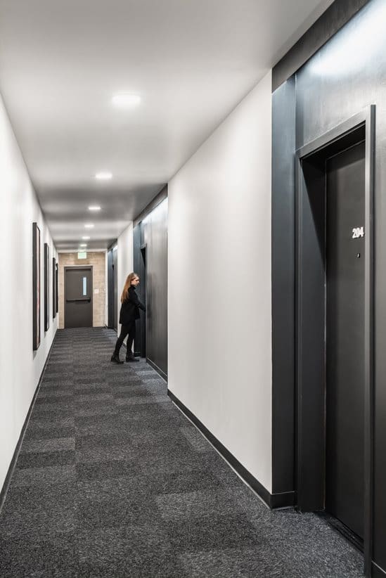 A long, narrow corridor with dark gray carpet tiles and white walls,